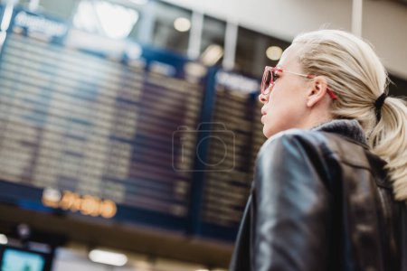 Téléchargez les photos : Jeune voyageuse élégante vêtue occasionnellement vérifiant un tableau des départs au terminal de l'aéroport en face de l'enregistrement dans les couteurs. Affichage de l'horaire de vol bleu en arrière-plan. Focus sur la femme
. - en image libre de droit