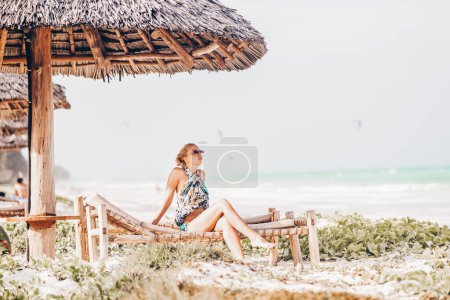 Téléchargez les photos : Femmes bronzant sur une chaise longue sous un parapluie en bois sur une magnifique plage tropicale. Kiteboarders lagon bleu turquoise de la plage de Paje, Zanzibar, Tanzanie en arrière-plan
. - en image libre de droit