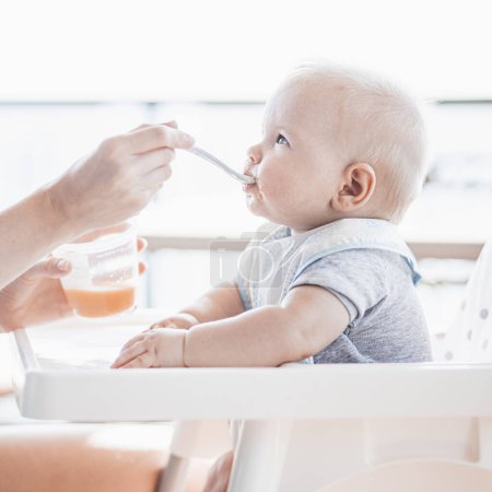 Photo for Mother spoon feeding her baby boy child in baby chair with fruit puree on a porch on summer vacations. Baby solid food introduction concept - Royalty Free Image
