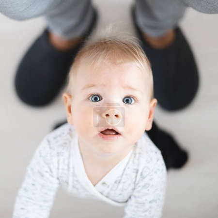 Téléchargez les photos : Vue du haut vers le bas de joyeux bébé garçon faisant les premiers pas tenant au pantalon de survêtement de pères à la maison. Bébé garçon mignon apprenant à marcher. - en image libre de droit