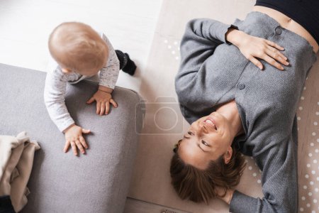 Photo for Happy family moments. Mother lying comfortably on childrens mat playing with her baby boy watching and suppervising his first steps. Positive human emotions, feelings, joy - Royalty Free Image