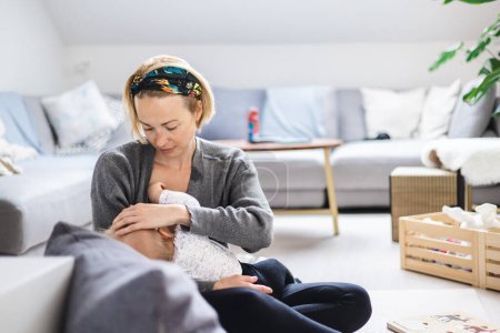 Foto de Mujer joven amamantando a su bebé bebé niño casualy sentado en los niños jugando estera en el piso de la sala de estar en casa - Imagen libre de derechos