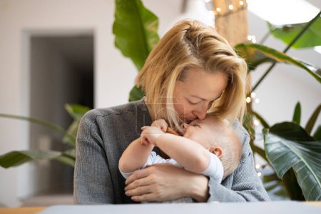 Photo for Portrait of young mother cuddling and kissing her adorable little baby boy while sitting at the table at home. Sensory stimulation for baby development - Royalty Free Image