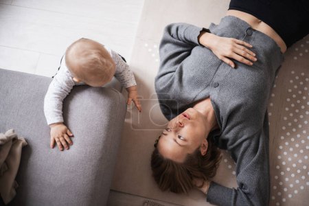 Photo for Happy family moments. Mother lying comfortably on childrens mat playing with her baby boy watching and suppervising his first steps. Positive human emotions, feelings, joy - Royalty Free Image
