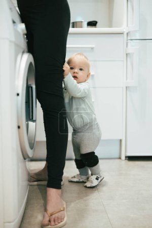 Téléchargez les photos : Little infant baby boy child hiding between mothers legs demanding her attention while she is multitasking, trying to do some household chores in kitchen at home. Mother on maternity leave - en image libre de droit