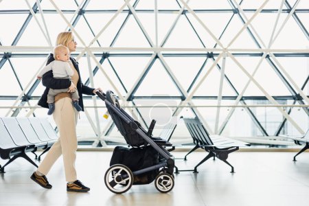 Photo for Mother carying his infant baby boy child, pushing stroller at airport departure terminal moving to boarding gates to board an airplane. Family travel with baby concept - Royalty Free Image