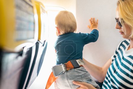 Téléchargez les photos : Maman et son enfant volent en avion. Mère tenant et jouant avec son bébé garçon sur ses genoux pendant le vol commercial de l'économie. Concept photo de voyage en avion avec bébé. De vraies personnes - en image libre de droit