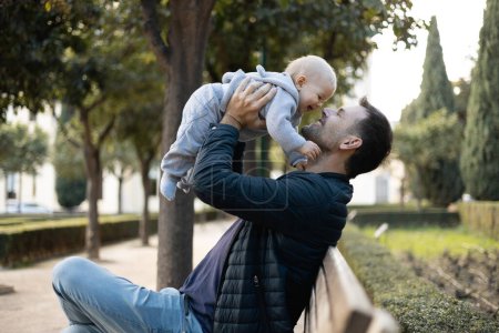 Father holding and lifting his cute infant baby boy child sitting on wooden bench in urban city park. Dad and son enjoing a pristine moment of happiness, smiling and laughing.