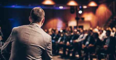 Ein Redner hält einen Vortrag über Unternehmenskonferenzen. Unerkennbare Menschen im Publikum im Konferenzsaal. Veranstaltung für Unternehmen und Unternehmertum.
