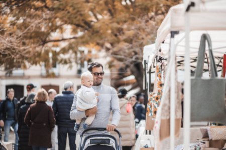Père marchant portant son bébé garçon enfant et poussant une poussette dans la foule de personnes visitant le marché aux puces du dimanche à Malaga, Espagne