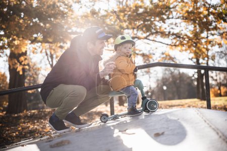 Téléchargez les photos : Père supervise son petit garçon courageux tout en roulant scooter bébé à l'extérieur dans le skate park urbain. Casque de protection jaune pour enfant. - en image libre de droit