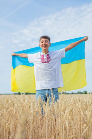 Téléchargez les photos : Priez pour l'Ukraine. Enfant avec drapeau ukrainien dans le champ de blé. Garçon souriant en vêtements ukrainiens nationaux avec jaune - grand drapeau bleu à la main sur fond de champ de blé. Éducation patriotique. - en image libre de droit