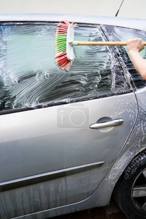 Kaukasische Männer waschen Scheiben schmutziger grauer Autos mit Bürste und weißer Seife, Autowäsche mit Bürste
