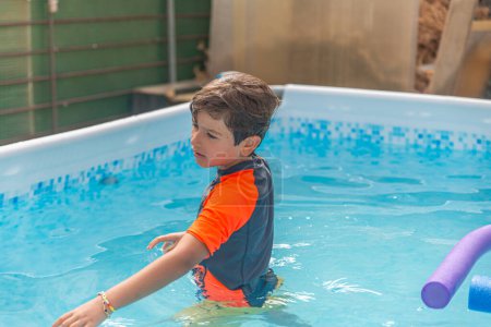 Nachdenklicher kleiner Junge in orangefarbenem Badehemd, der in einem Pool nachdenklich wegschaut