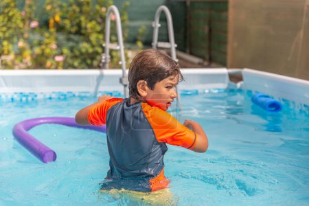 A focused young boy wades through the water in a pool, a purple noodle floating nearby, amidst vibrant greenery