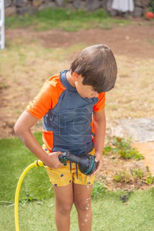 Niño curioso en camisa naranja examinando manguera de jardín al aire libre, con salpicaduras de agua visibles, día soleado.