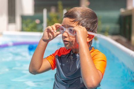 Thoughtful child in swim gear looks contemplatively away in a sunny backyard pool.