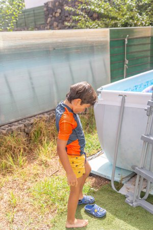 Niño pequeño y pensativo vestido con vibrante traje de baño de pie junto a una escalera de piscina de metal, contemplando tomar un refrescante baño en un día de verano brillante y soleado.