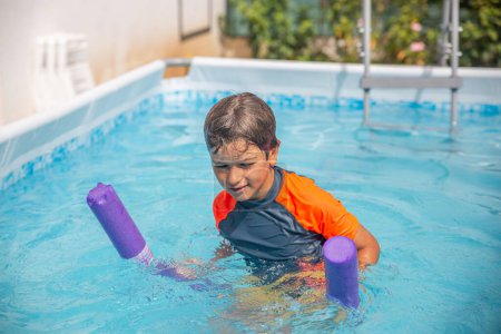 Kleines Kind im Pool mit einer lila Nudel, um an einem hellen und sonnigen Tag schwimmen zu lernen