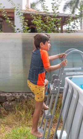 Really young boy in an orange shirt and colorful life vest climbs the pool ladder, looking away in thought.
