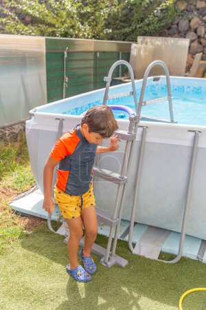 Pequeño joven limpiando el agua de su cara después de disfrutar del tiempo de juego en una piscina del patio trasero, de pie pensativamente al lado de la escalera de la piscina en un día soleado cálido.