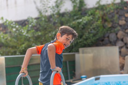 Joyful smiling boy in a bright swim shirt turning with a swim noodle in the sparkling backyard pool.