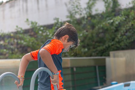 Niño en traje de baño de verano pensativamente de pie junto a la piscina, mirando hacia abajo