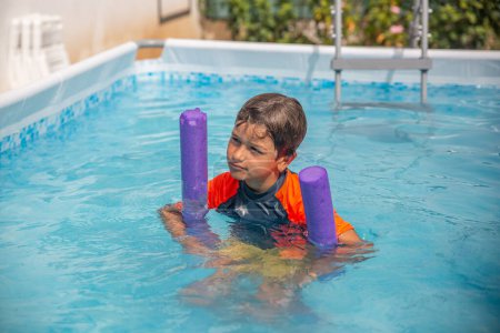 Garçon heureux avec nouilles de natation pourpres profitant de l'eau dans une piscine extérieure, excité pour le plaisir d'été