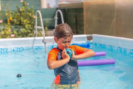 Fröhlicher Junge im heimischen Pool spielt mit Wasserball, mit Schwimmnudel und üppigem Garten im Hintergrund
