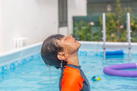 Boy in orange swim shirt enjoys a cool off, head tilted back with water cascading, noodles, and pool stairs