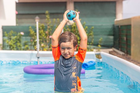 Boy exudes joy holding a ball aloft in a swimming pool, marking a moment of pure fun in the sun