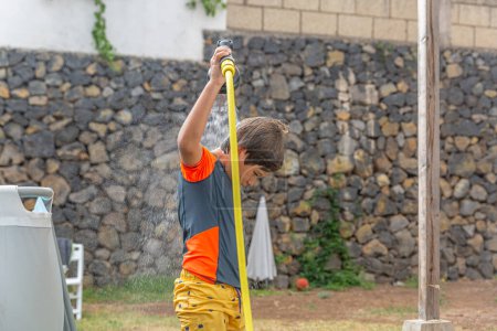 Photo for Seen from behind, a child demonstrates control and power over a garden hose, actively spraying water high, forming a sparkling cascade of droplets during a dynamic play session. - Royalty Free Image