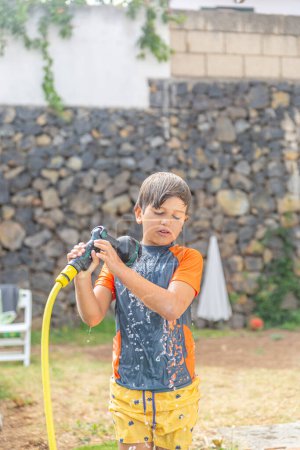 Young boy in wet sports attire handling garden hose with a focused expression in a sunny backyard.