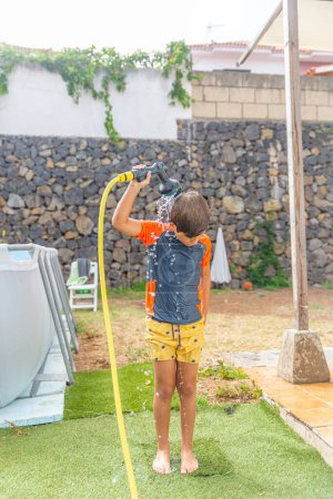 Boy in garden playfully splashing water from hose over head, enjoying summer with stone wall background.
