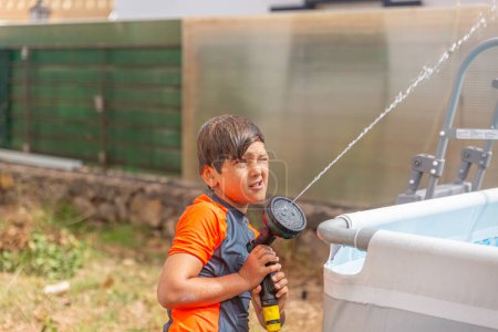 Young boy in orange shirt squinting against sunlight while spraying water from a hose.