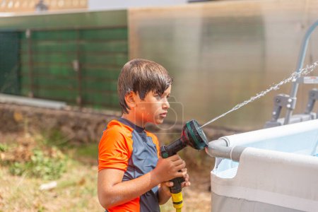 Thoughtful boy in sportswear watering with garden hose, focusing on water jet, clear day.