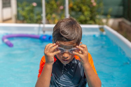 Young child carefully adjusting swimming goggles poolside, preparing to dive into clear blue pool water.