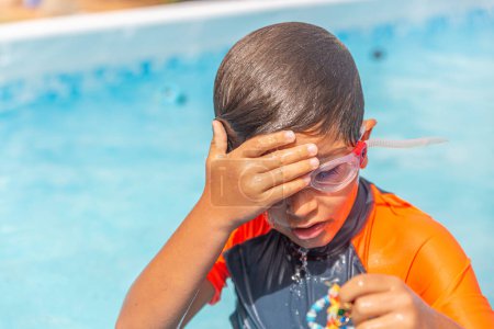 Young child at the poolside wiping water away from eyes, goggles lifted after a refreshing swim.