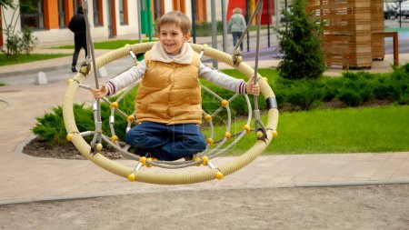 Foto de Movimiento lento de niño feliz divirtiéndose balanceándose en el columpio de cuerda en el patio público. Niños activos, deportes y desarrollo, niños jugando al aire libre. - Imagen libre de derechos