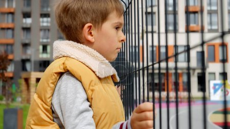 Téléchargez les photos : Triste petit garçon tenant une clôture métallique sur une aire de jeux publique et regardant à travers. Dépression infantile, problèmes d'intimidation, victimes à l'école, émigration, criminalité et pauvreté. - en image libre de droit
