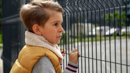 Photo for Portrait of lonely boy holding hands on metal fence and looking through it. Child depression, problems with bullying, victim in school, emigration, criminal and poverty. - Royalty Free Image