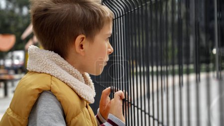 Foto de Niño sintiéndose triste y solo mirando a través de la valla de metal en el parque infantil. Depresión infantil, problemas con el acoso escolar, víctima en la escuela, emigración, delincuencia y pobreza. - Imagen libre de derechos