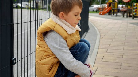 Photo for Little upset boy sitting next to metal fence feeling unhappy and lonely. Child depression, problems with bullying, victim in school, emigration, criminal and poverty. - Royalty Free Image
