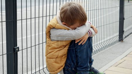 Photo for Lonely boy feeling alone on playground sitting next to metal fence and crying. Child depression, problems with bullying, victim in school, emigration, criminal and poverty. - Royalty Free Image