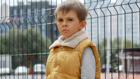 Photo for Upset, sad and lonely boy leaning on metal fence at public playground. Child depression, problems with bullying, victim in school, emigration, criminal and poverty. - Royalty Free Image