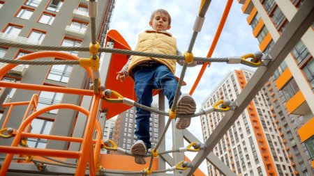 Foto de Pequeño chico activo caminando sobre el puente de cuerda en el parque infantil moderno. - Imagen libre de derechos