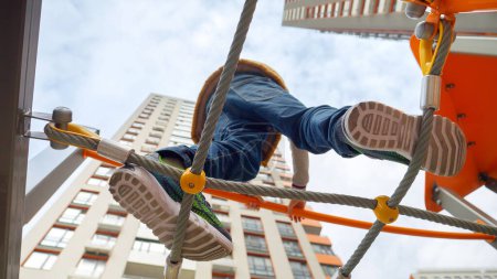 Foto de Vista desde el suelo en el niño en zapatillas de deporte de pie en la torre alta en el patio de recreo y walkng sobre el puente de cuerda. - Imagen libre de derechos