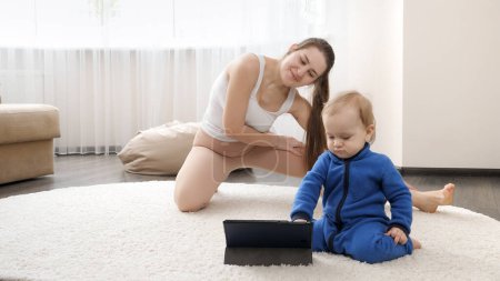 Photo for Young mother with little baby son doing sports on carpet and watching training video on tablet computer. Family healthcare, active lifestyle, parenting and child development. - Royalty Free Image