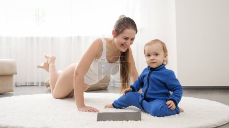 Photo for Cheerful smiling baby boy holding tablet computer while mother is doing push-ups and fitness exercises on carpet. Family healthcare, active lifestyle, parenting and child development. - Royalty Free Image