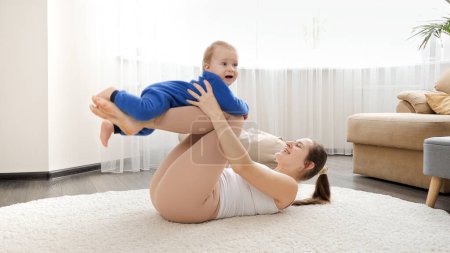 Téléchargez les photos : Jeune femme couchée sur le sol et berçant son fils bébé tout en faisant des exercices de fitness. - en image libre de droit